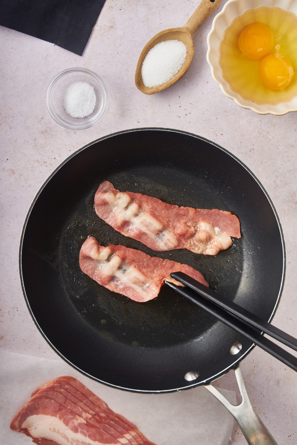 A black skillet with two pieces of bacon cooking and one piece being flipped over using a pair of black tongs.