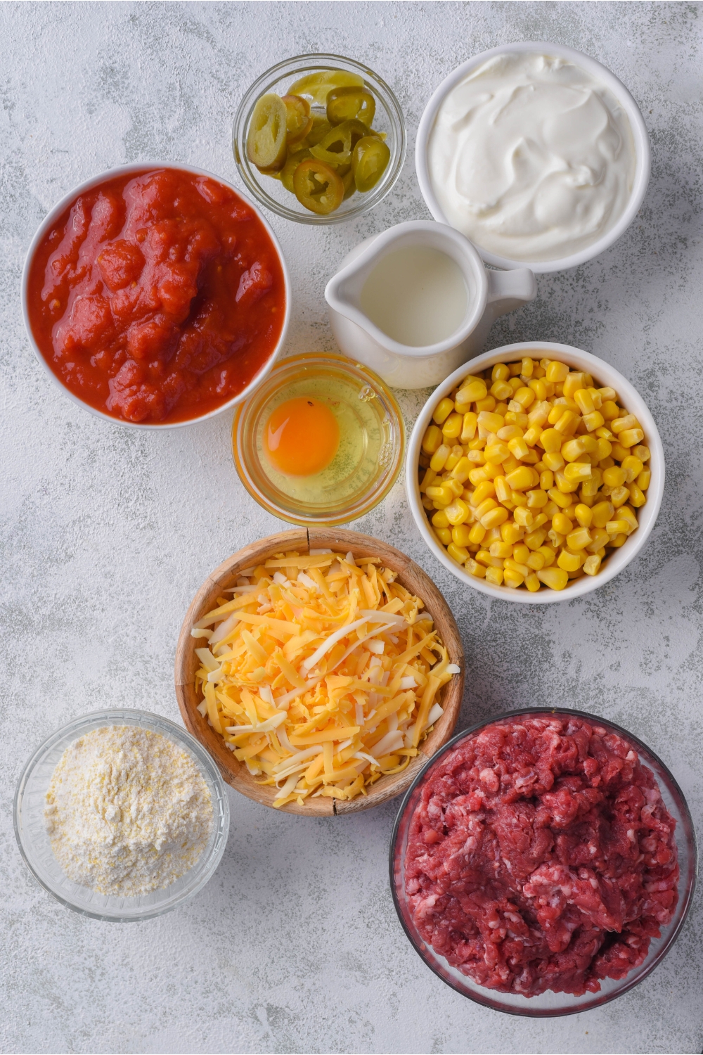 Overhead view of an assortment of ingredients including bowls of sour cream, an egg, cornbread mix, crushed tomatoes, corn, raw ground beef, and shredded cheese.