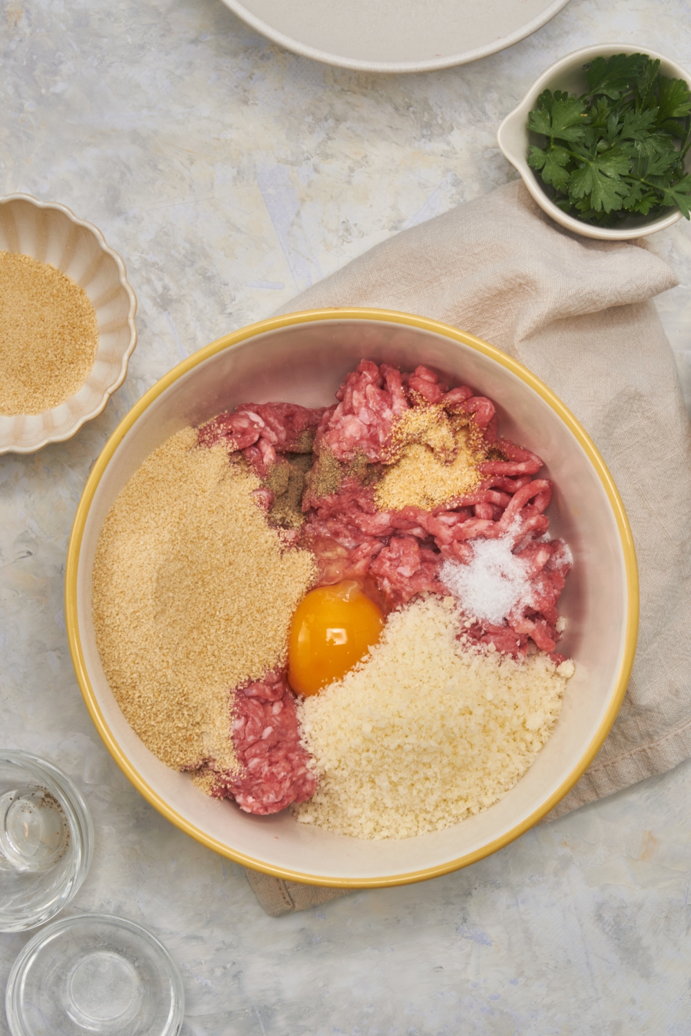 Overhead view of a white and yellow bowl filled with raw ground beef, bread crumbs, parmesan cheese, an egg, and spices. None of the ingredients have been combined.