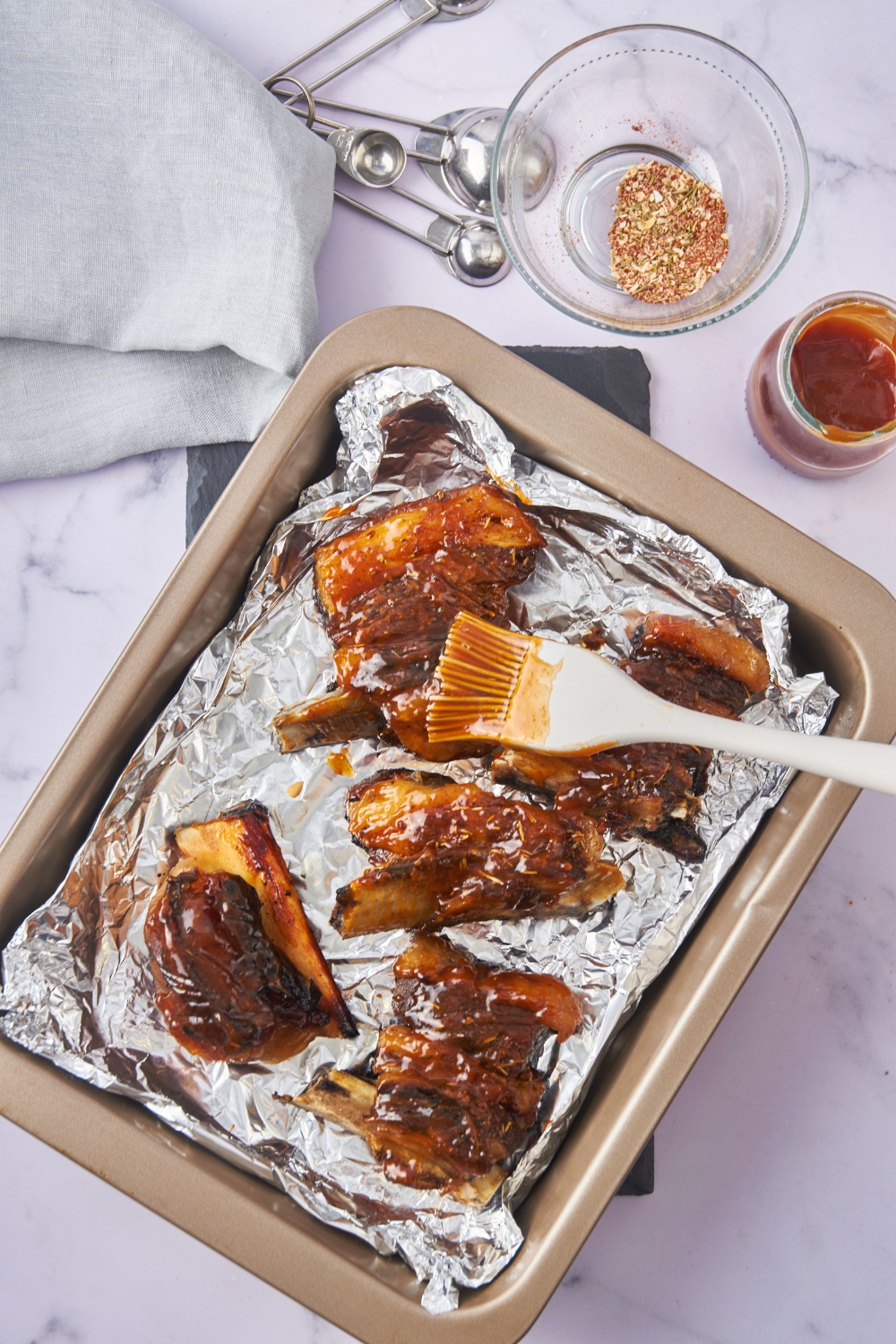 Five beef back ribs coated in BBQ sauce spread evenly in a baking dish lined with foil and a silicone basting brush is brushing sauce on the ribs.