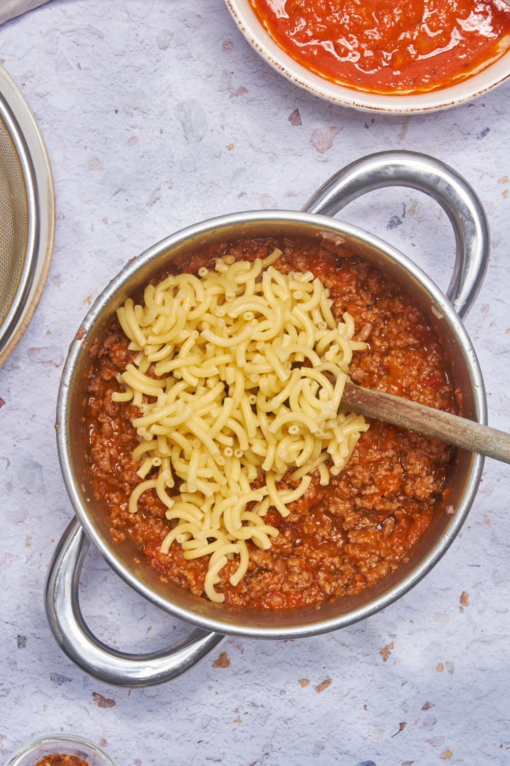 A large pot with two handles filled with a tomato and meat sauce and cooked macaroni noodles freshly added to the pot.