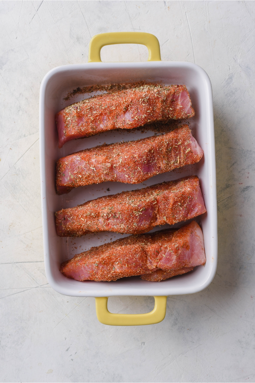 Overhead view of four seasoned and boneless ribs placed evenly in a white baking dish with yellow handles.