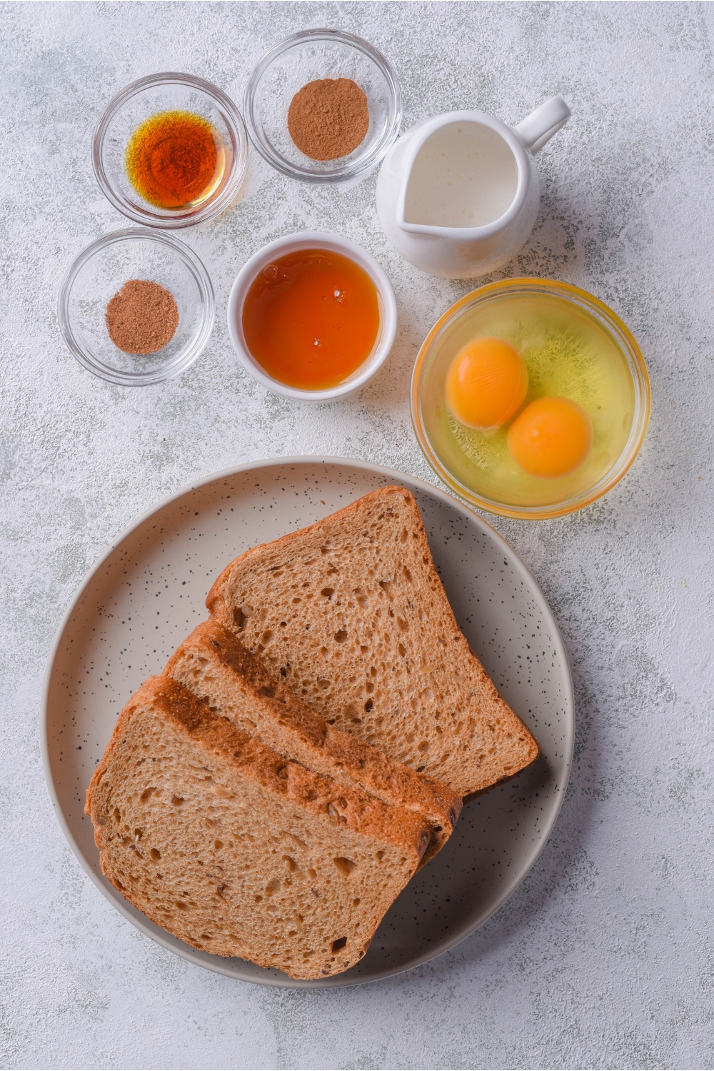 An assortment of ingredients including a plate of sandwich bread and bowls of eggs, cream, vanilla extract, cinnamon, and maple syrup.