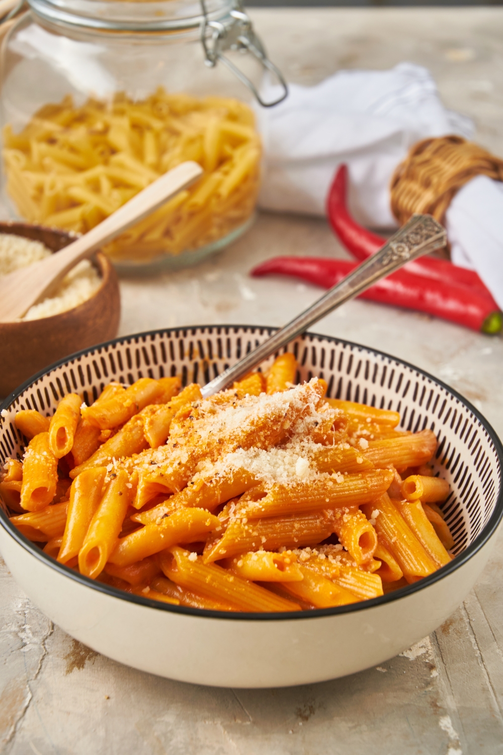 A bowl of Carbone spicy rigatoni garnished with grated parmesan cheese and in the background is a jar of dried pasta and a bowl of parmesan cheese with a fork in it.