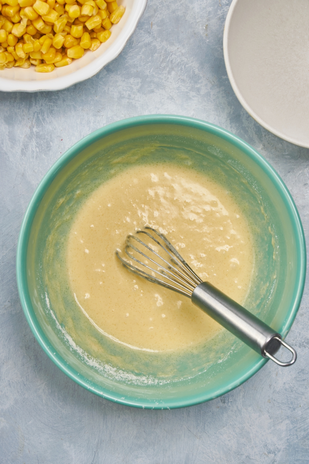 A blue bowl filled with cornbread batter and a whisk is in the bowl.