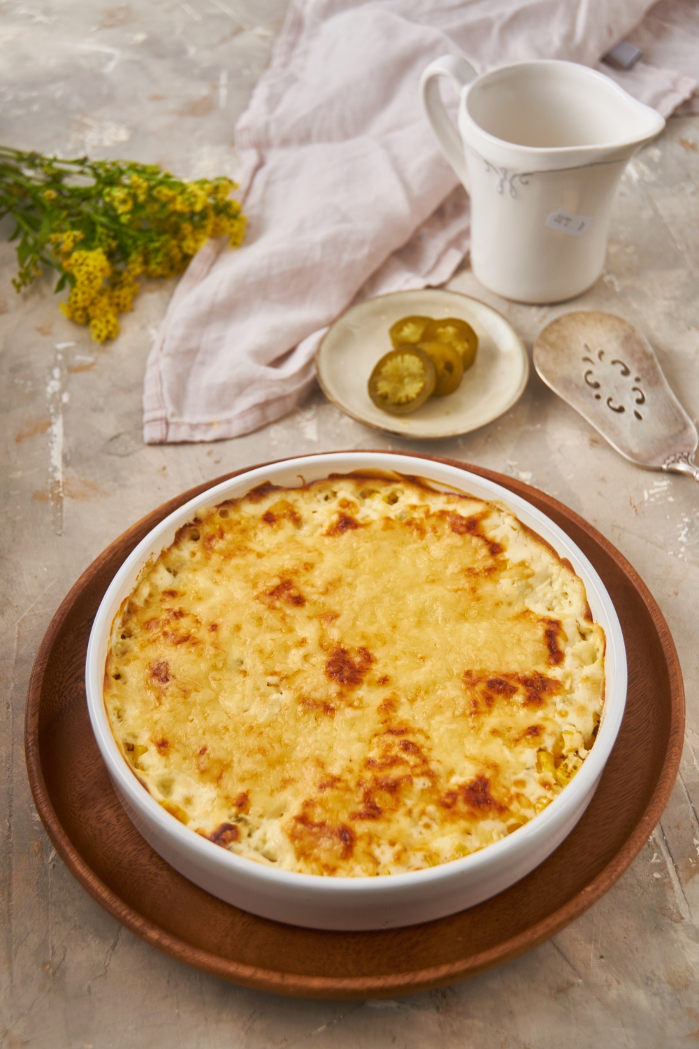 A white round baking dish filled with baked cream cheese corn casserole. The casserole is on top of a wooden plate.