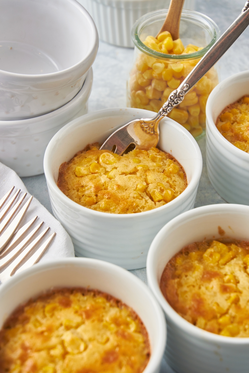 White ramekins filled with golden brown corn casserole. There is a fork in one of the ramekins and a jar of corn is next to it.