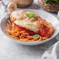 A bowl of chicken parmesan with chicken covered in melted cheese, a garnish of fresh basil, and served on a bed of pasta with marinara sauce.