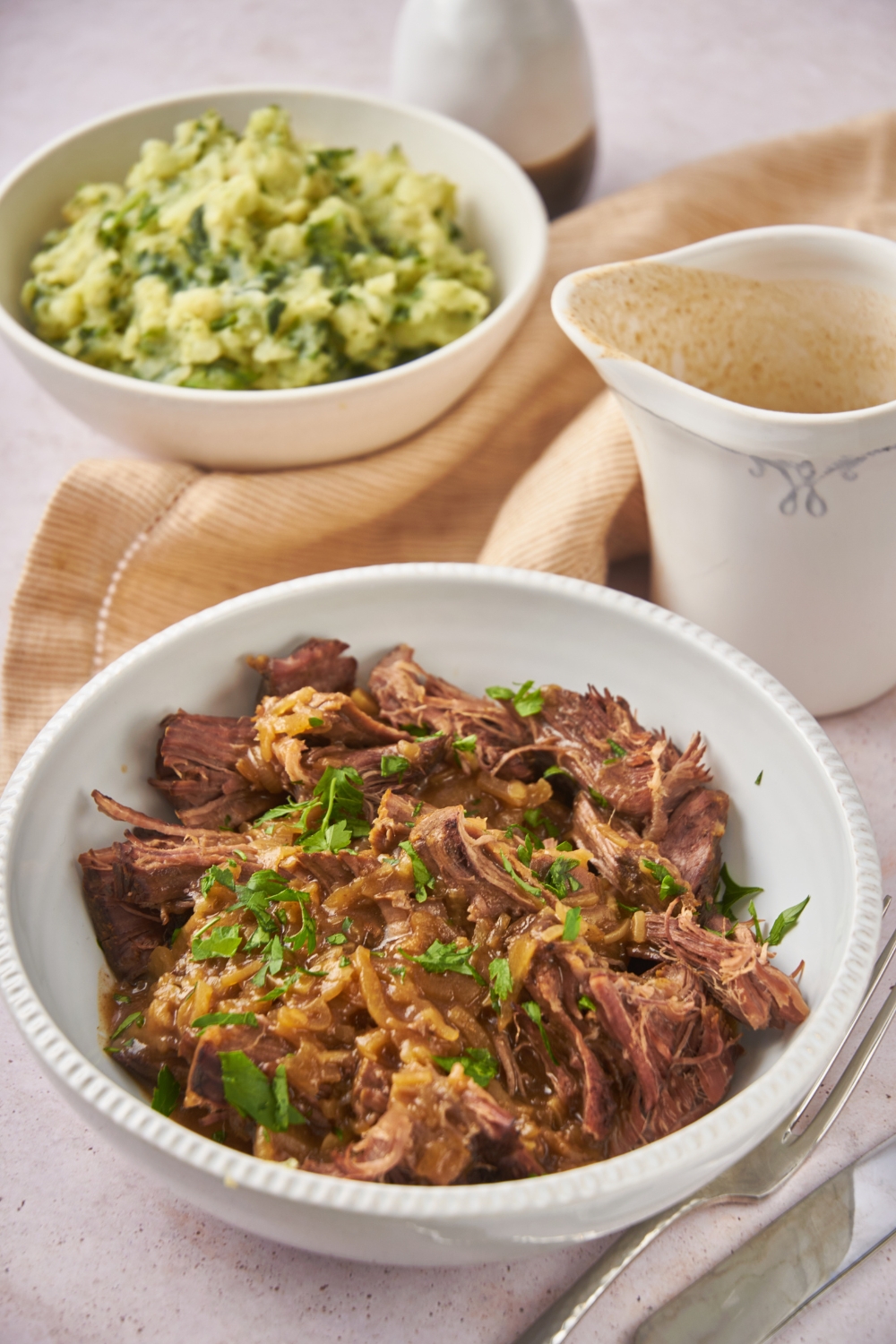 A bowl of cooked and shredded eye of round beef covered in a thin brown sauce and garnished with fresh green herbs.