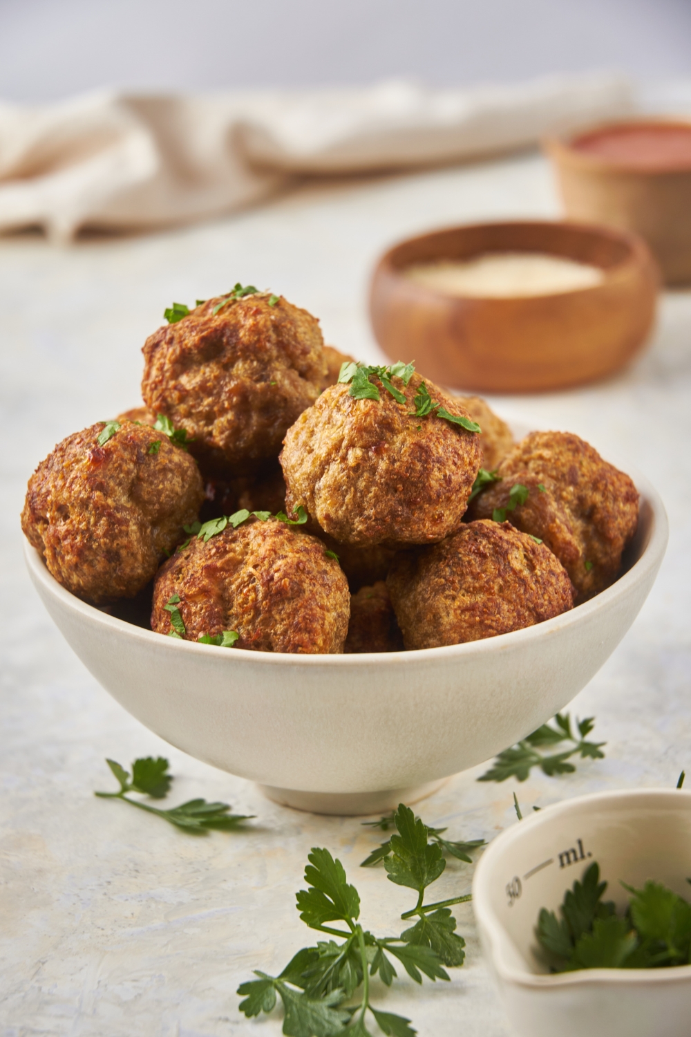 A bowl filled with baked meatballs that have been garnished with fresh green herbs, some of which are sprinkled around the counter.