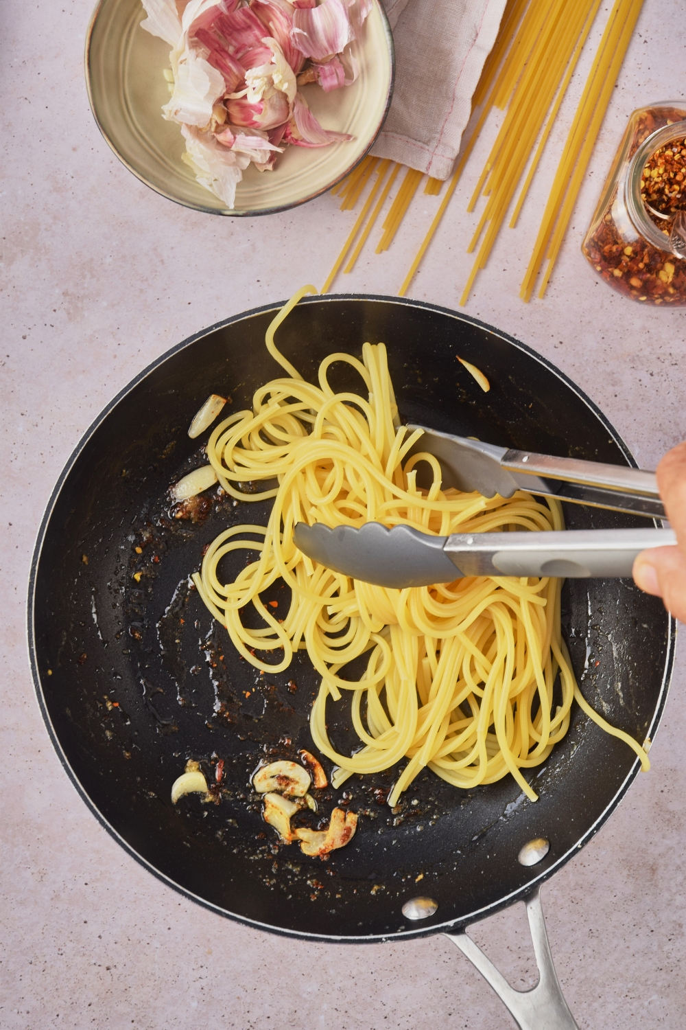 Black skillet with oil, garlic, and red pepper flakes and spaghetti noodles being added to it using a pair of silicone tongs.