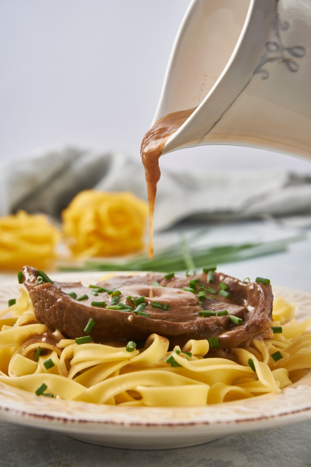 A container of gravy being poured over a cube steak covered in gravy with a bed of linguini noodles beneath it. Gravy is spilling from the container onto the steak.