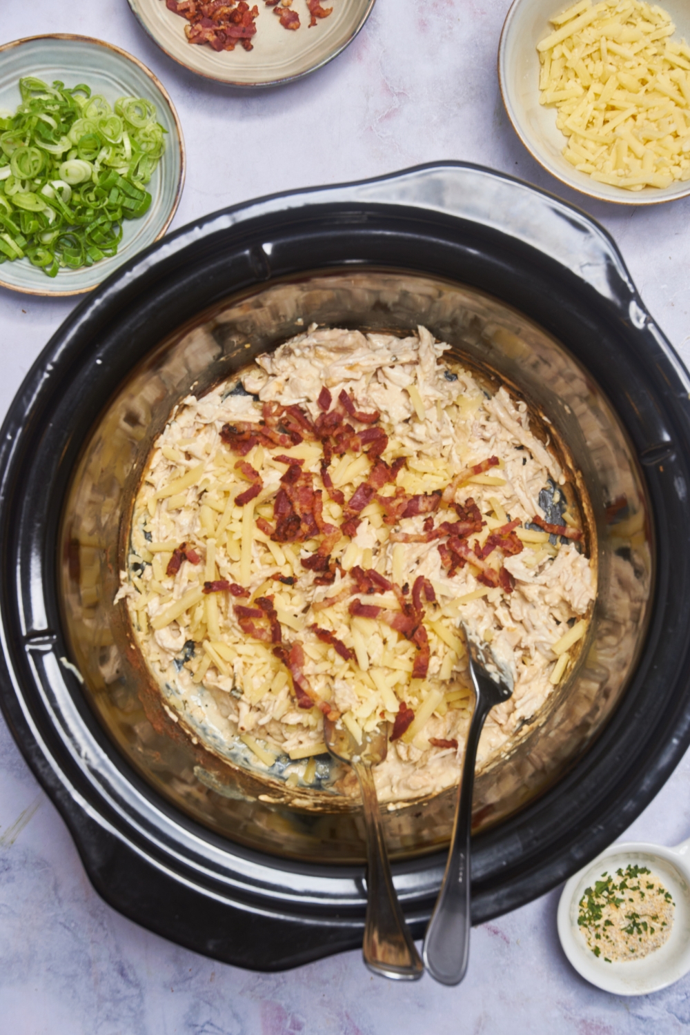 A crockpot filled with cooked and shredded chicken in a cream sauce, shredded cheese, bacon, and two forks in it.