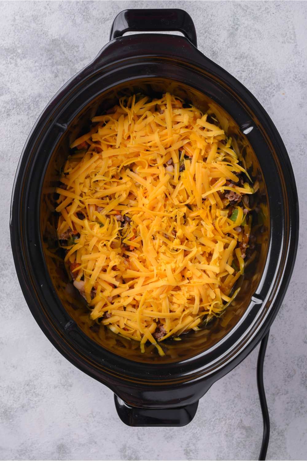 A black slow cooker with casserole and a layer of shredded cheddar cheese on top.