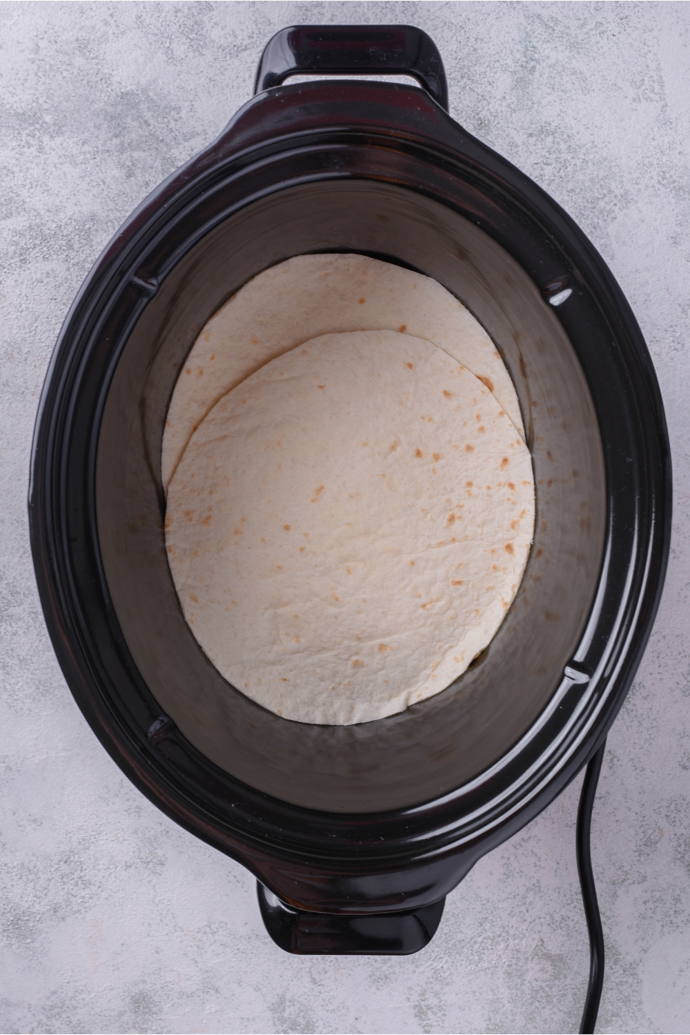 A black slow cooker with two tortillas on the bottom.