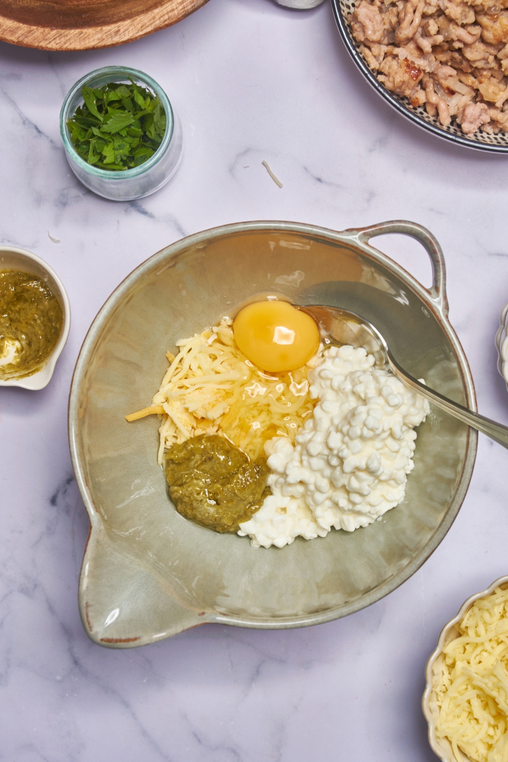 A mixing bowl filled with ricotta cheese, pesto, an unbeaten egg, shredded cheese, and a spoon.