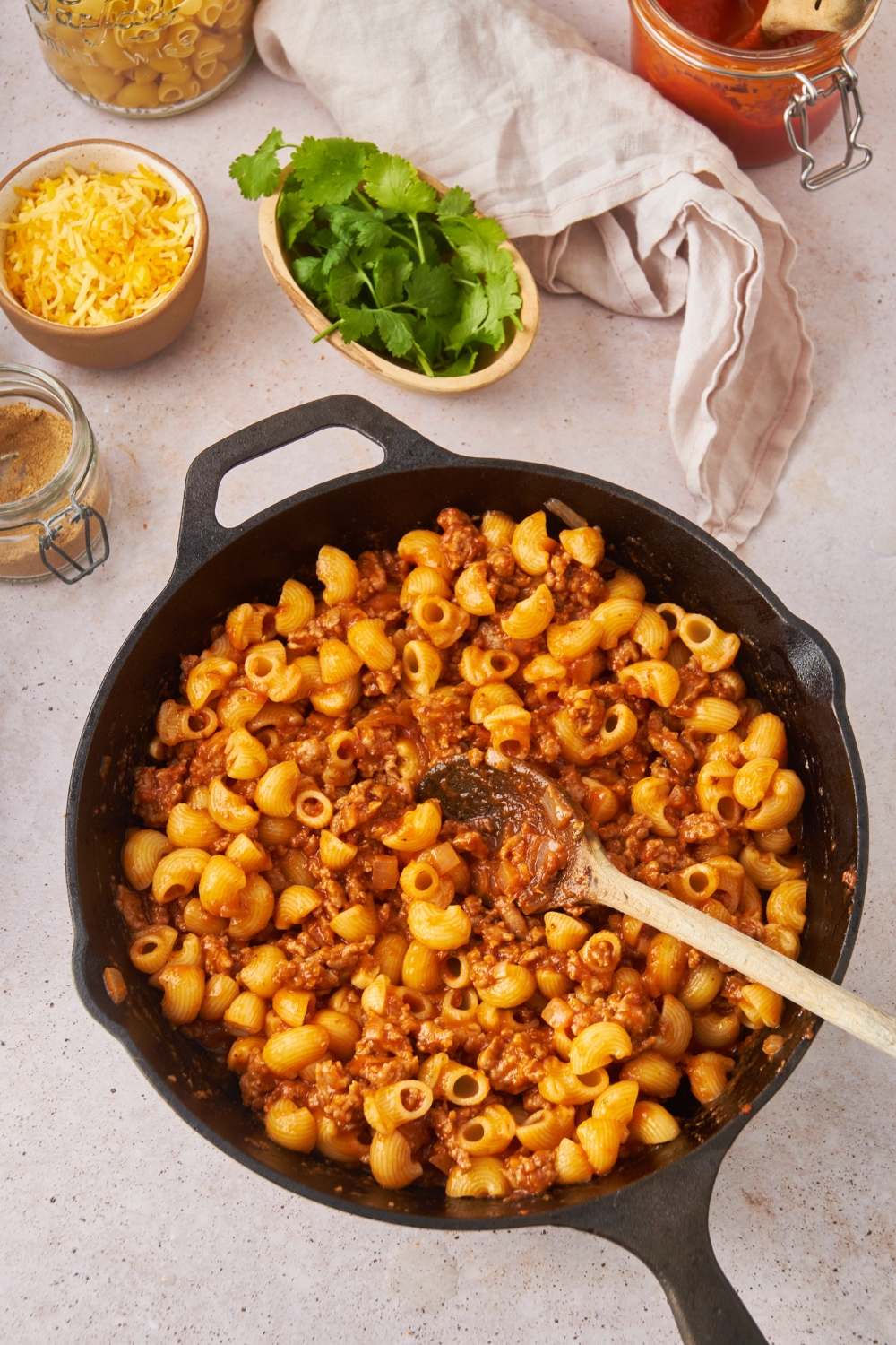 A black cast iron skillet with pasta, ground beef, and salsa combined in it using a wooden spoon.