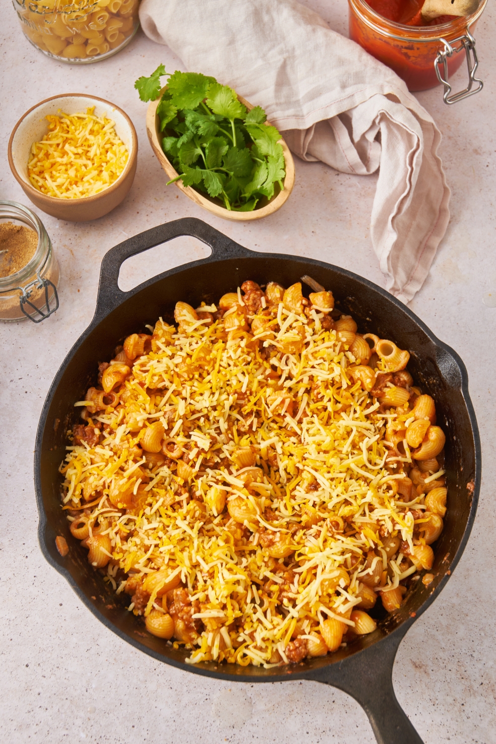 A black cast iron skillet with an unbaked taco pasta casserole covered in shredded cheese.