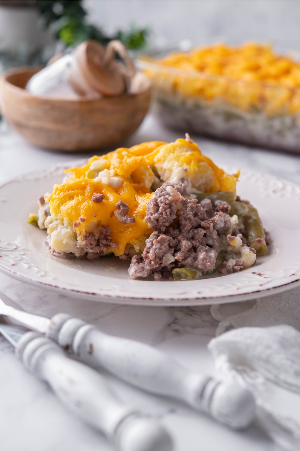 A serving of casserole with tater tots, green beans, ground beef, and shredded cheese on a white plate with the rest of the casserole in the background.