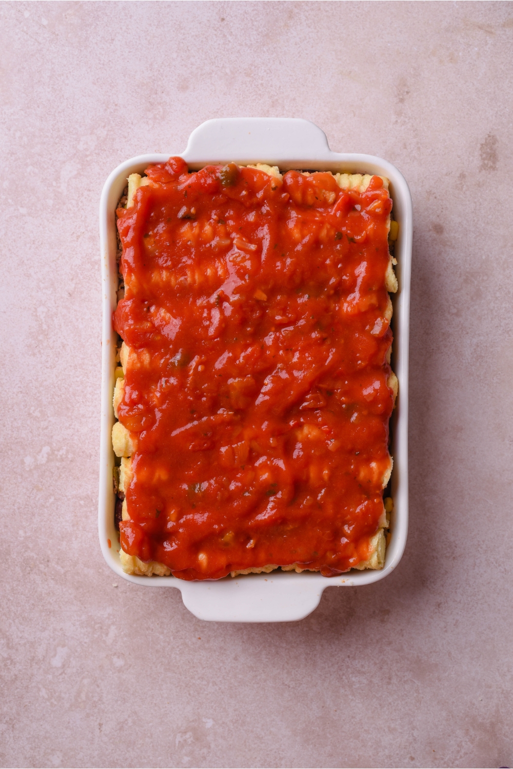 A white casserole dish filled with unbaked casserole, a layer of tater tots, and enchilada sauce poured over top.