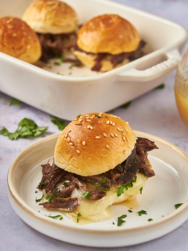 A mini tri tip sandwich filled with shredded beef, melted cheese, fresh green herbs, and topped with a poppyseed bun. In the background are three more tri tip sandwiches.