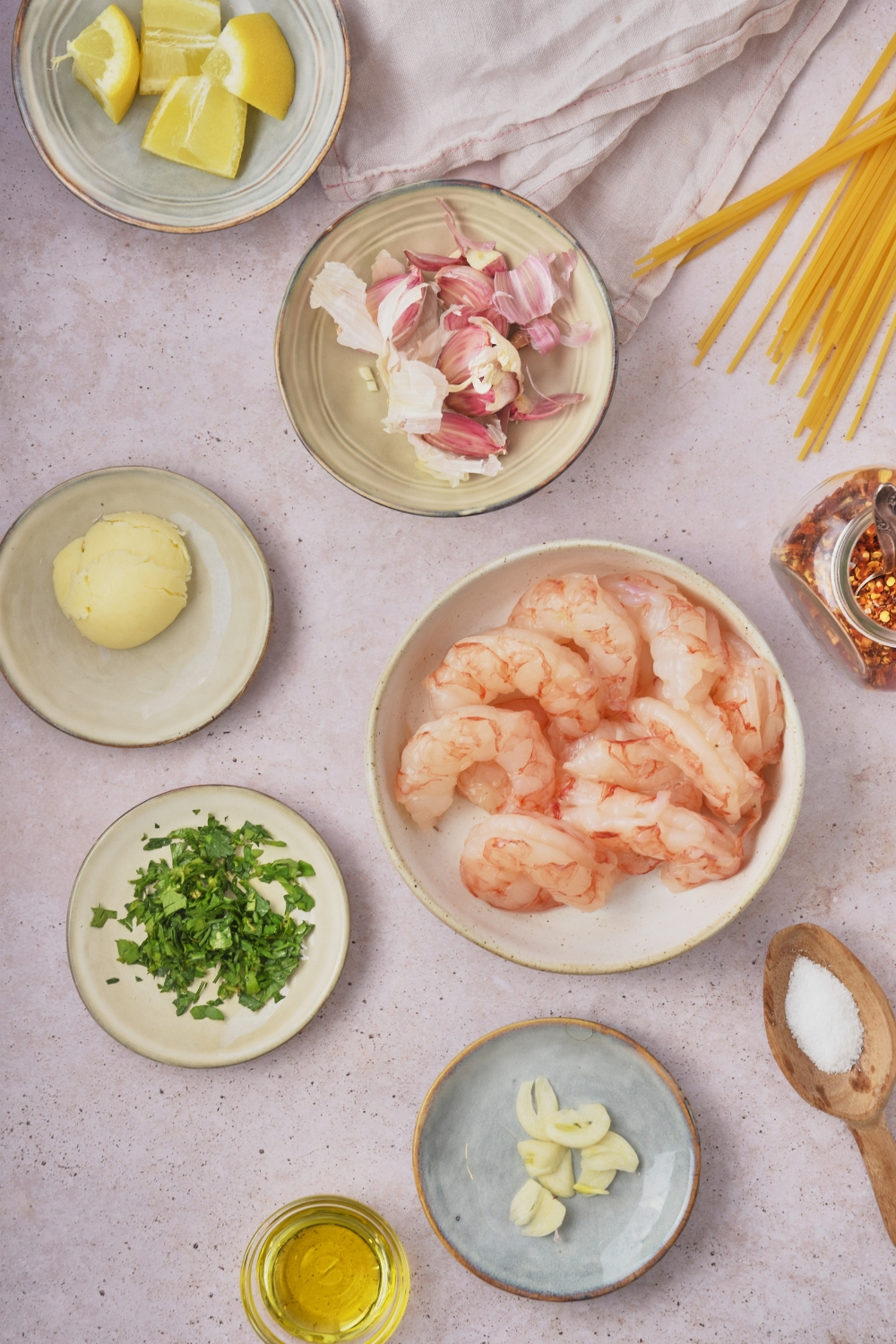 Overhead view of an assortment of ingredients including bowls of raw shrimp, butter, fresh green herbs, oil, garlic, and lemon slices.