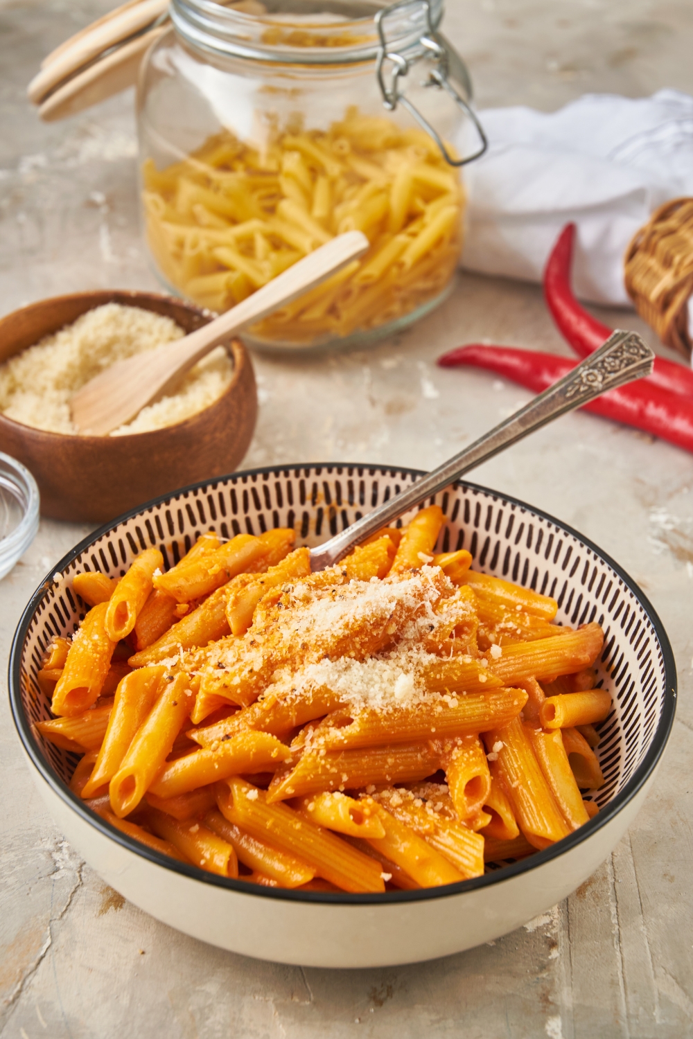 A bowl of Carbone spicy rigatoni garnished with grated parmesan cheese and in the background is a jar of dried pasta and a bowl of parmesan cheese with a spoon in it.