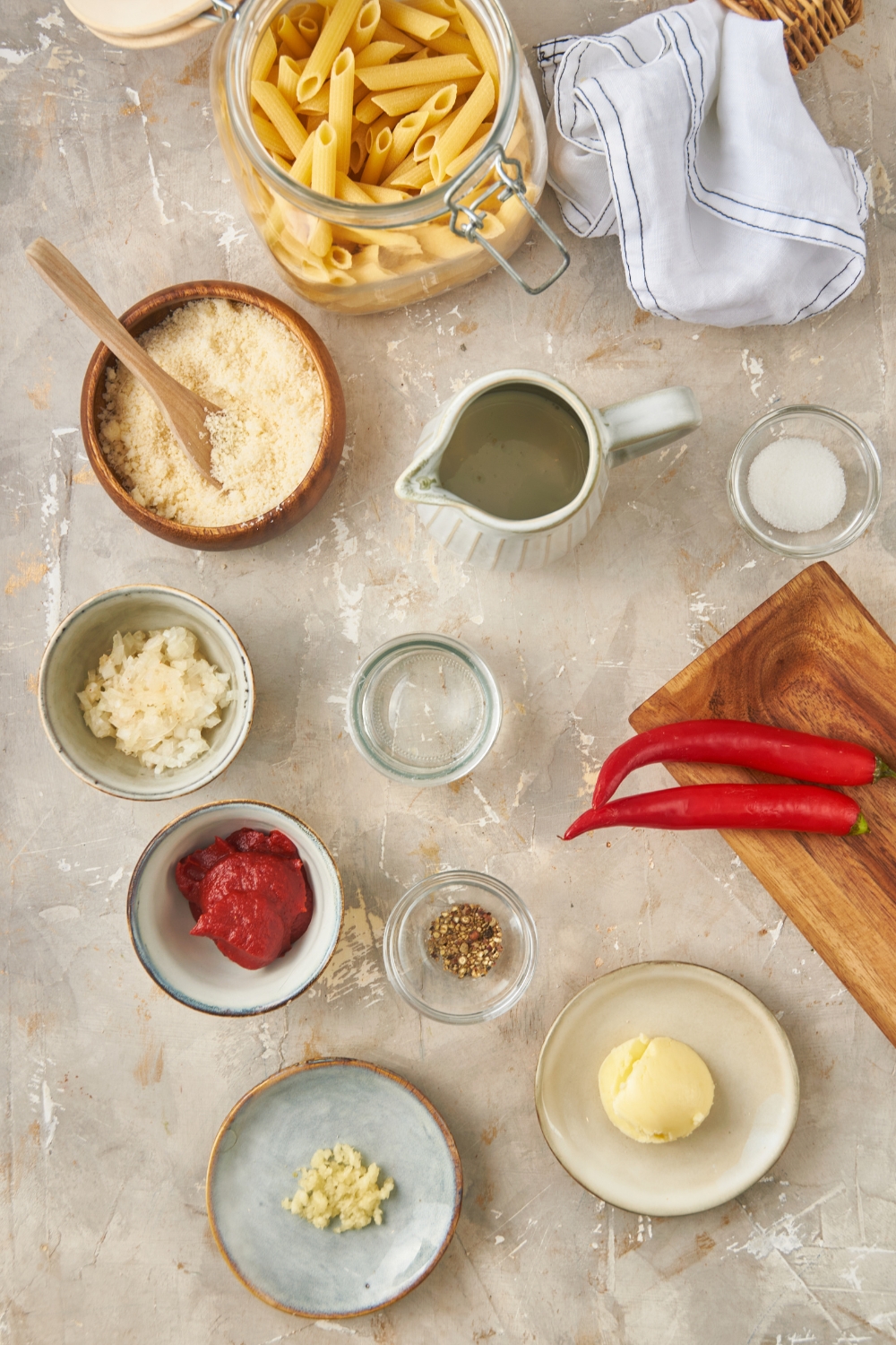 Overhead view of an assortment of ingredients including bowls of parmesan cheese, diced onion, tomato paste, a pitcher of vodka, two red chili peppers, a jar of dried pasta, a plate of minced garlic, and a plate of butter.