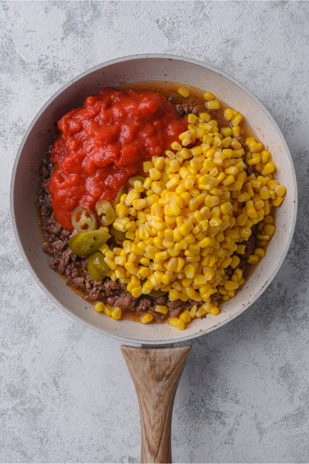 A grey skillet with cooked ground beef on the bottom and crushed tomatoes, corn, and jalapeños on top.