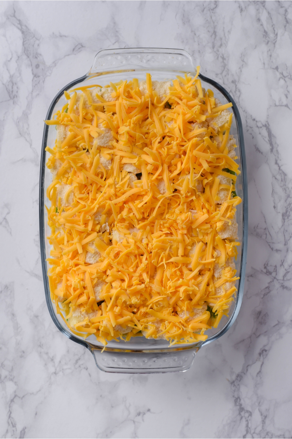 A clear baking dish filled with unbaked tater tot and green bean casserole that's covered in shredded cheddar cheese.