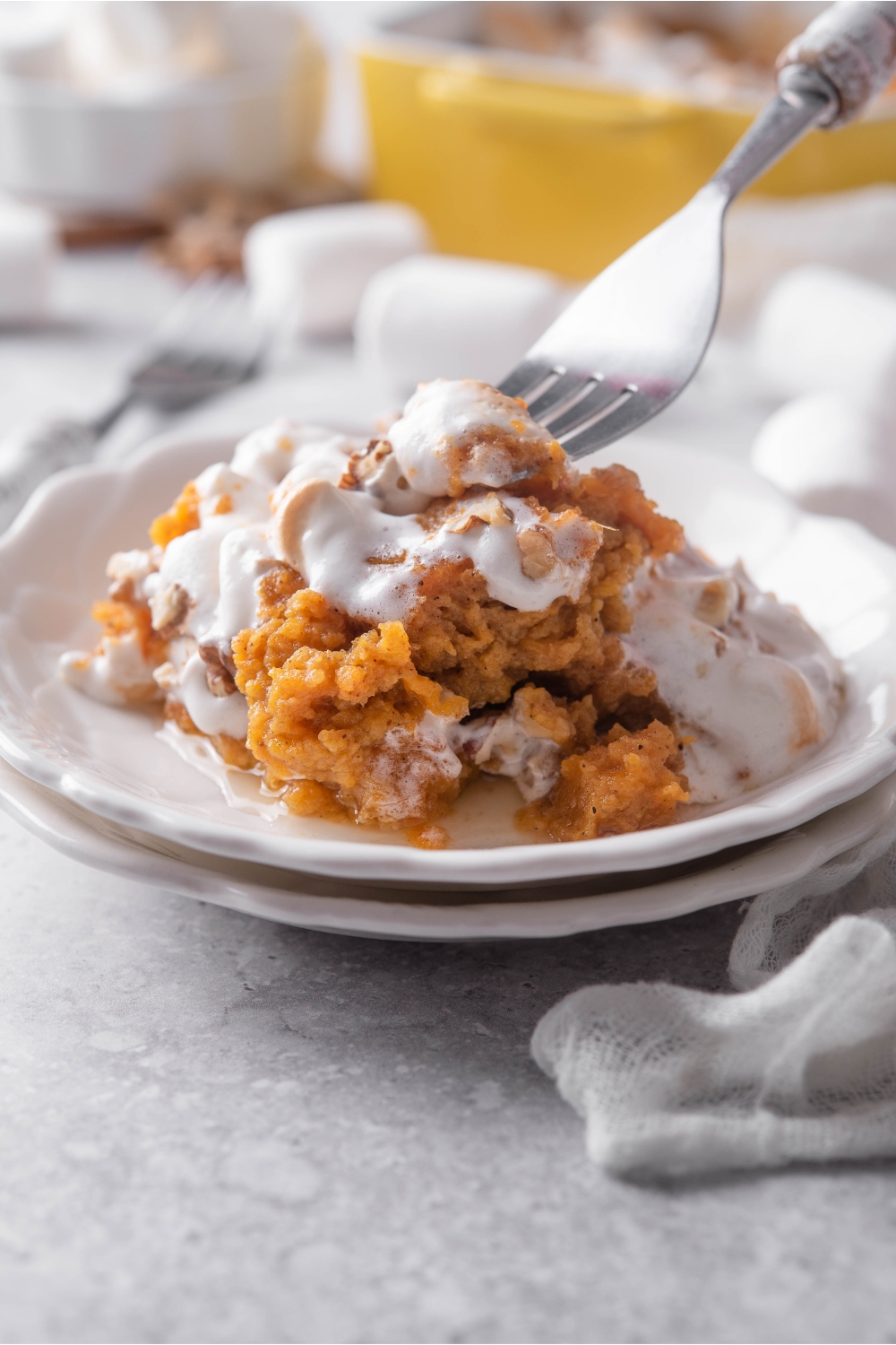 A serving of sweet potato casserole with marshmallows melting over top of the casserole. A fork is holding a piece of casserole over top of the plate.
