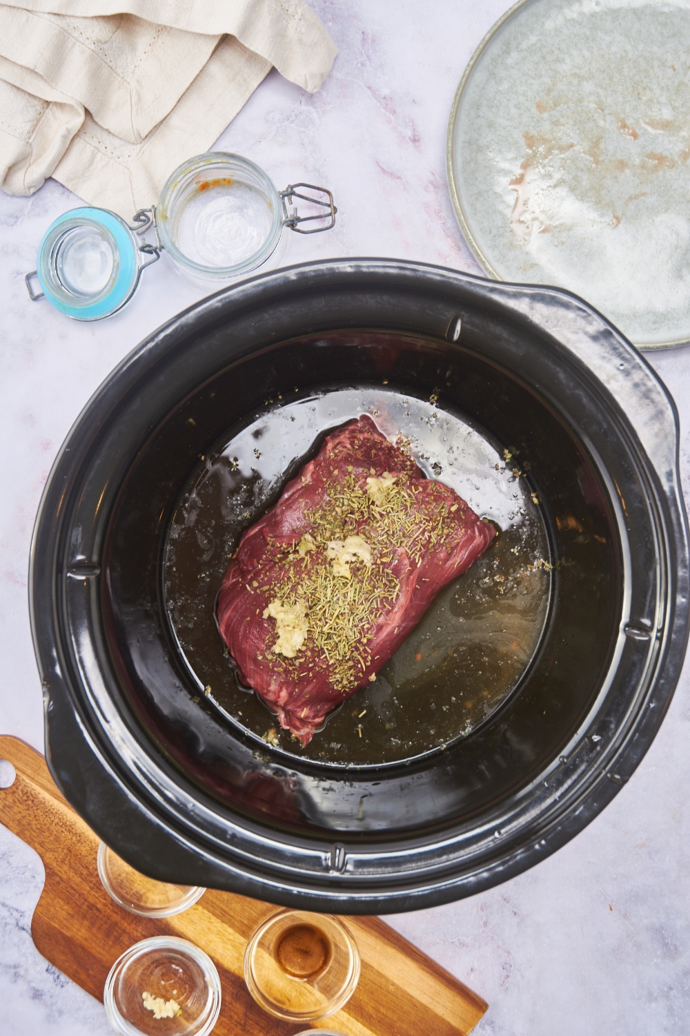 A raw beef roast in a black crock pot topped with minced garlic, dried herbs, and broth poured around the beef.