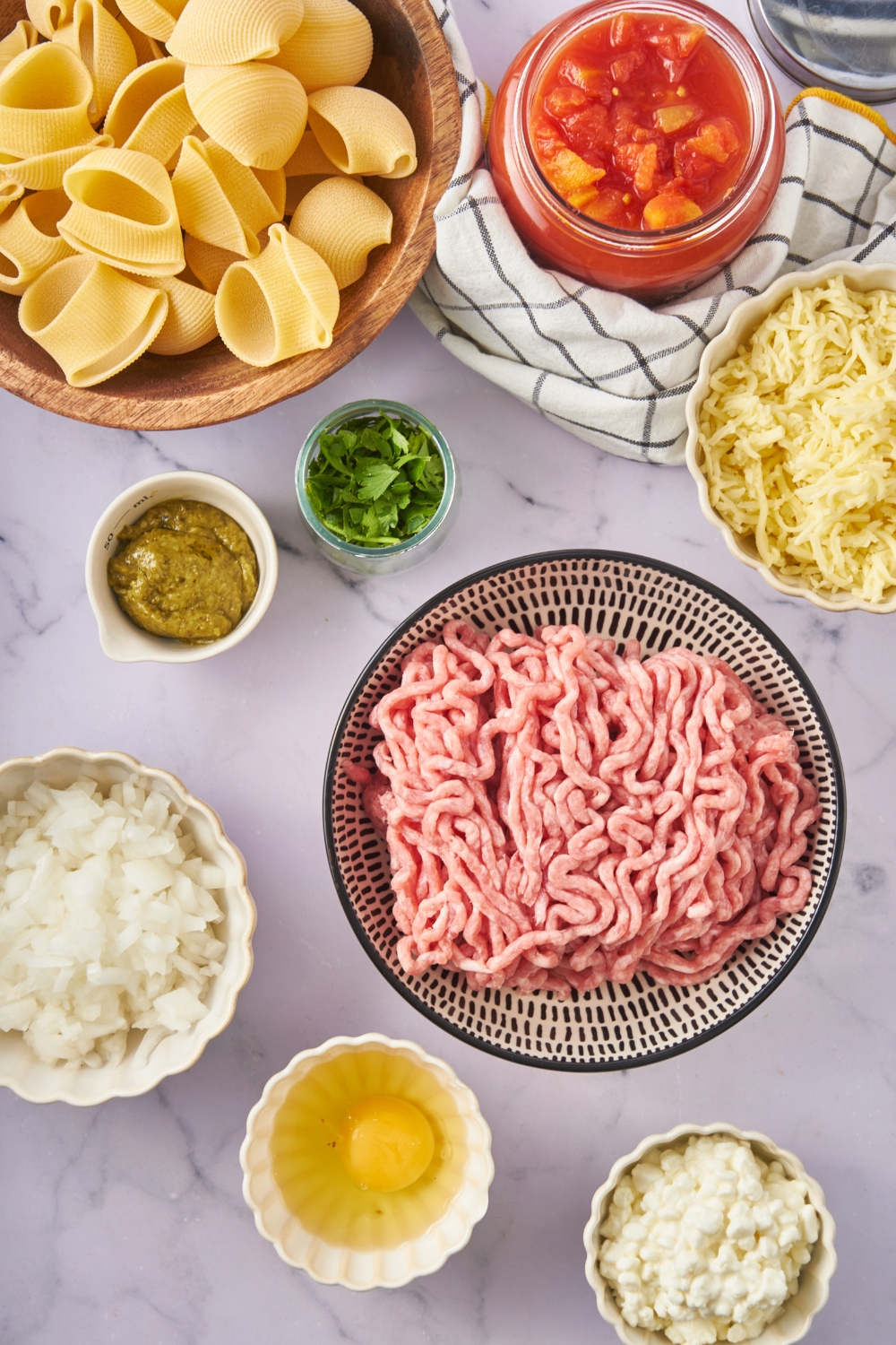 Overhead view of an assortment of ingredients including bowls of ground meat, tomato sauce, fresh herbs, pesto, dried pasta shells, diced onion, ricotta, and an unbeaten egg.