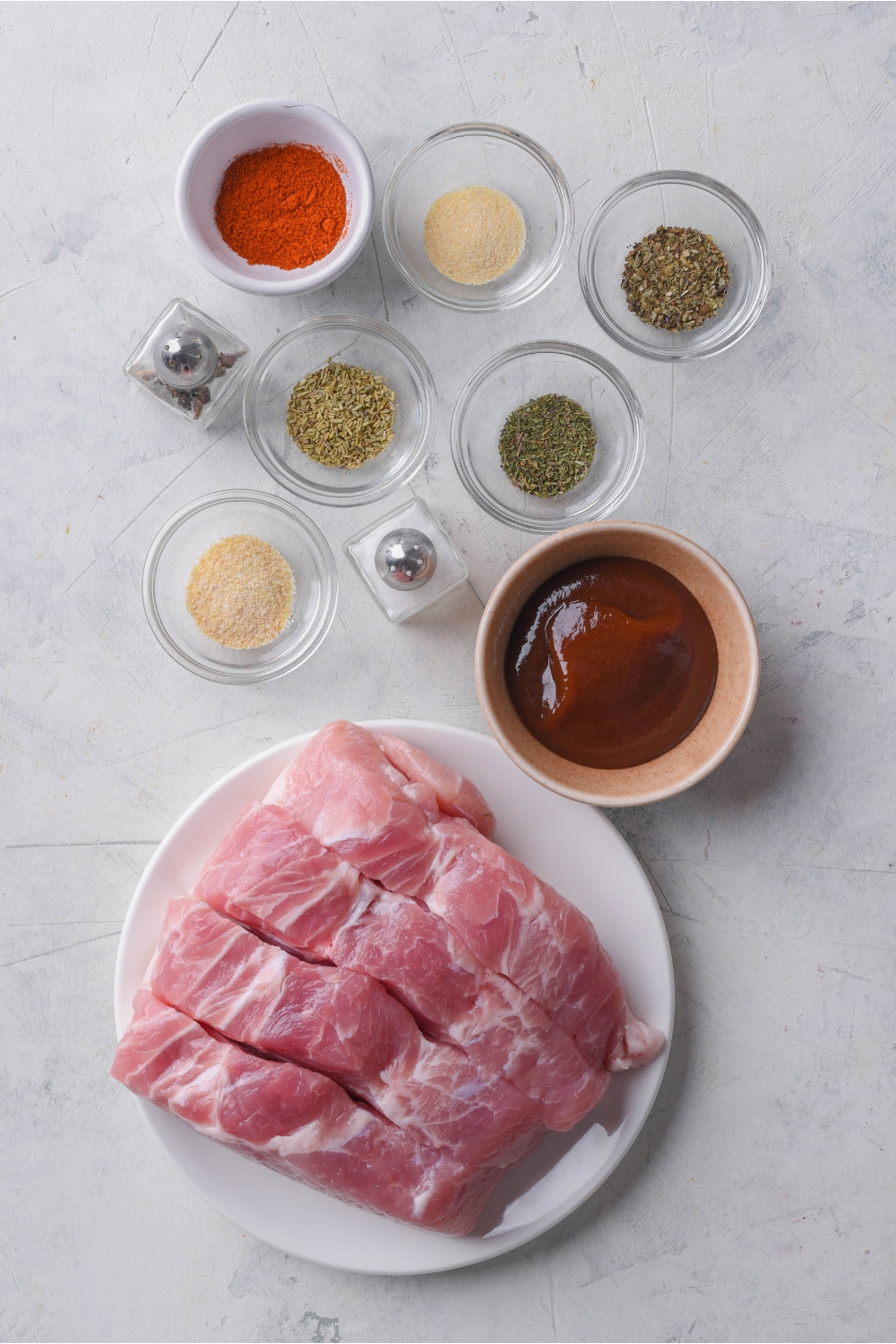 Overhead view of an assortment of ingredients including a plate of boneless pork ribs and bowls of barbecue sauce, various seasonings, and mini salt and pepper shakers.