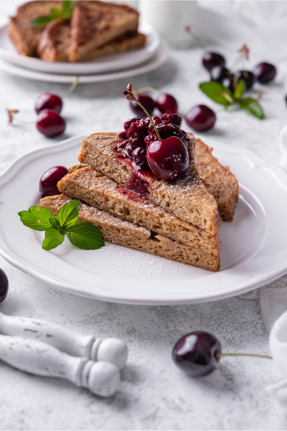 A stack of three pieces of French toast with a fourth piece behind the stack. The French toast is topped with cherry compote, a single cherry, and a mint sprig is garnished on the plate.