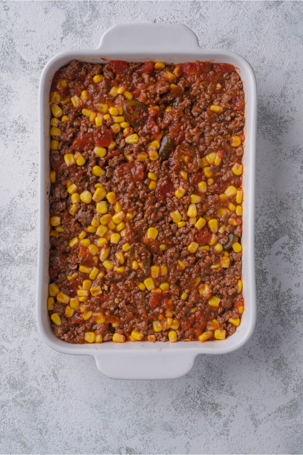 A white casserole dish with seasoned ground beef, corn, jalapeños, and crushed tomatoes in it.