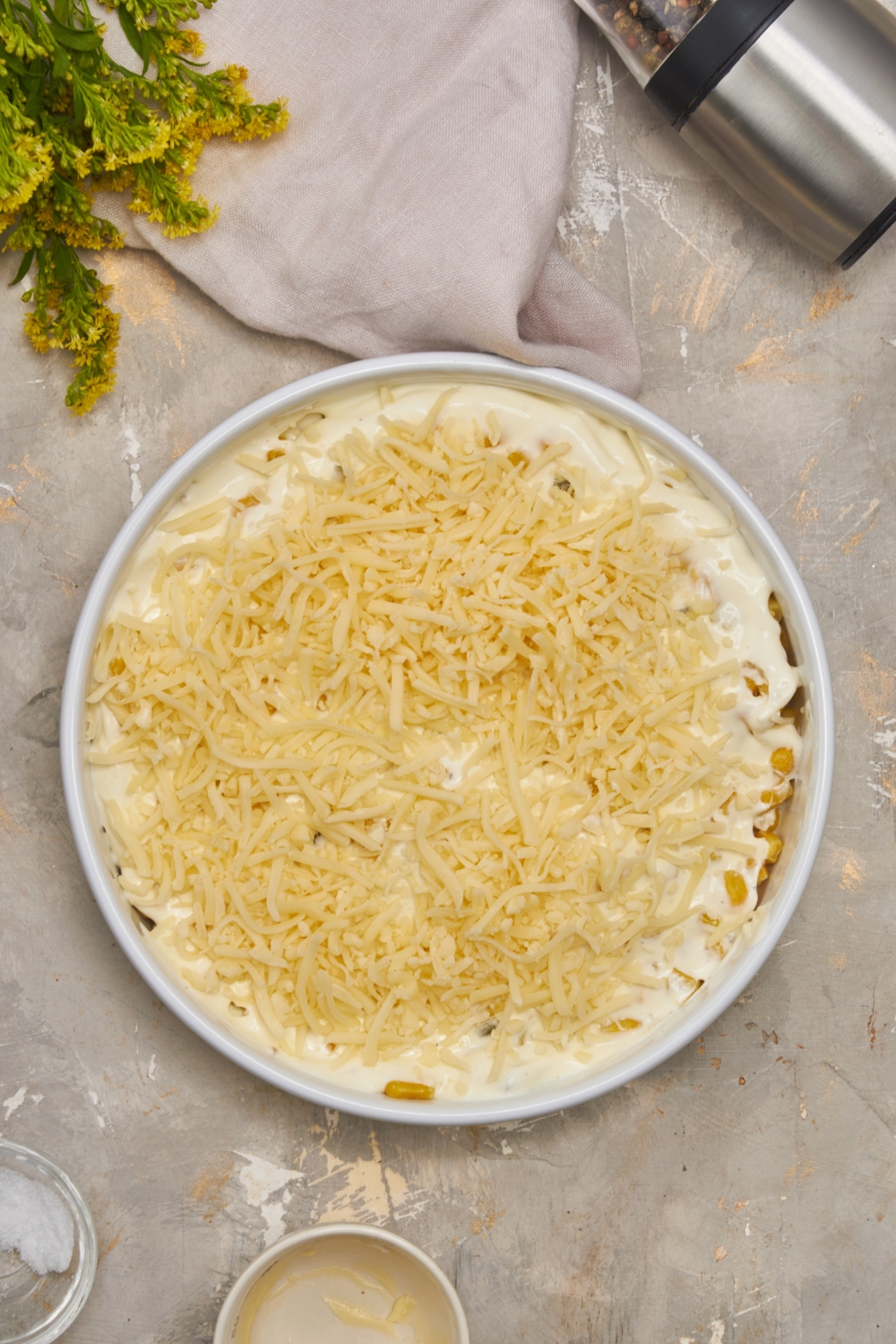 A white round baking dish filled with unbaked corn casserole and a pile of shredded cheese sprinkled on top of it.