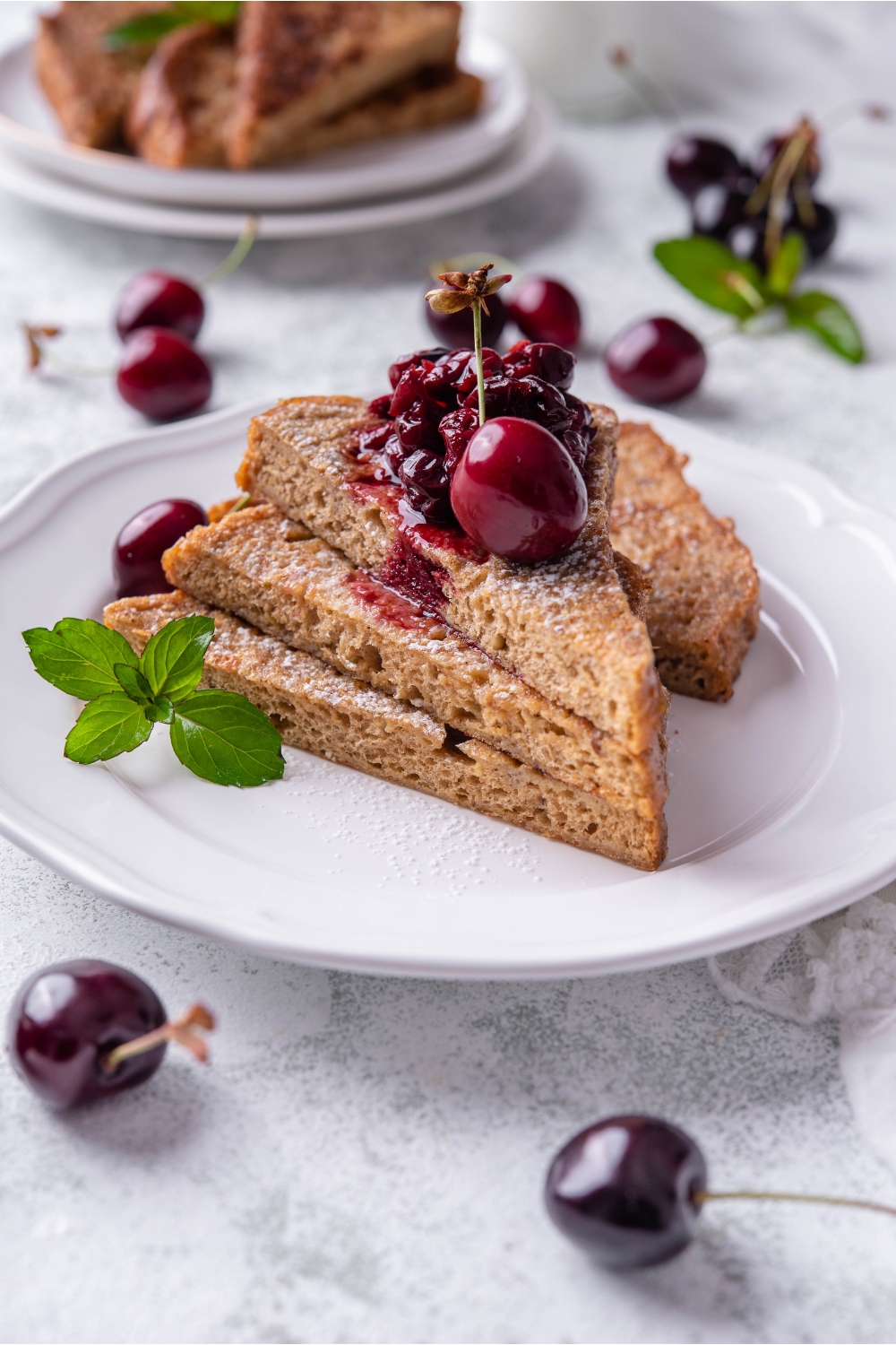 A stack of three pieces of French toast with a fourth piece behind the stack. The French toast is topped with cherry compote, a single cherry, and a mint sprig is garnished on the plate.