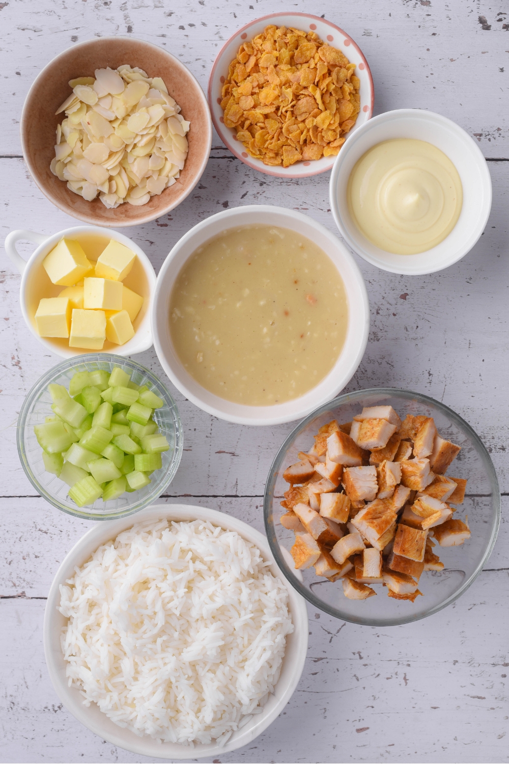Overhead view of an assortment of ingredients including bowls of cornflakes, chopped chicken, white rice, mayonnaise, slivered almonds, condensed chicken soup, and butter cubes.