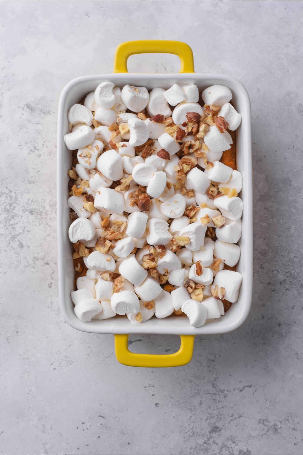 A white and yellow baking dish with sweet potato casserole, unbaked marshmallows, and chopped pecans sprinkled over top of the sweet potatoes.