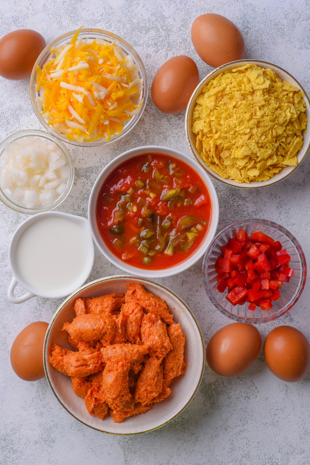 A countertop with multiple bowls of ingredients to make Mexican casserole. Eggs, diced tomatoes, diced bell pepper, chorizo, milk, diced onions, shredded cheese, and crushed chips are all on the counter.