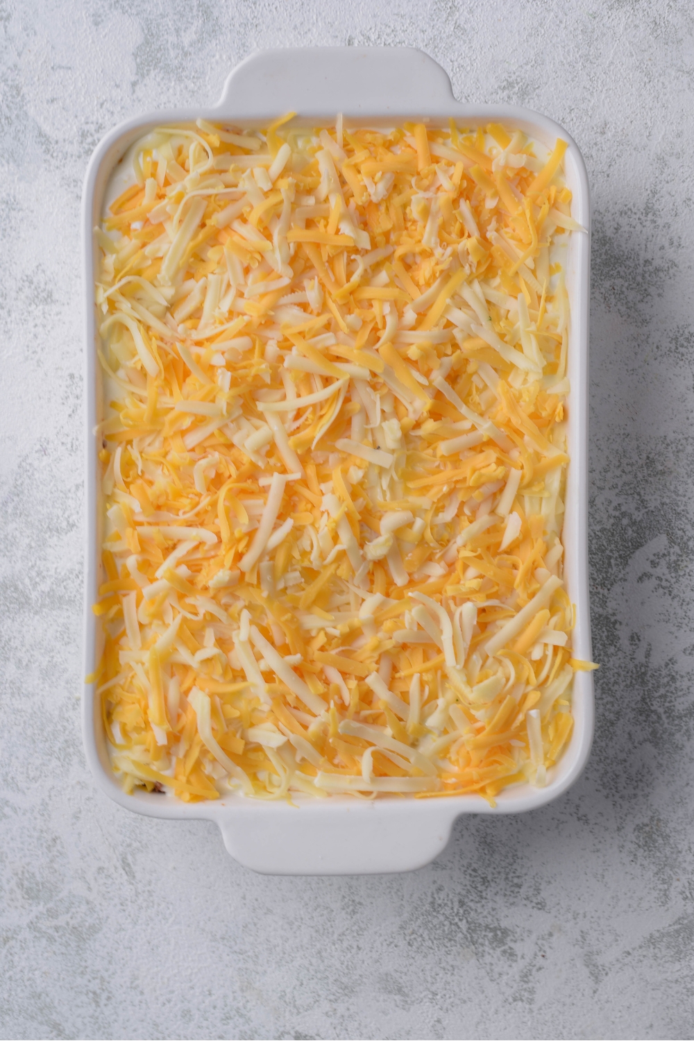 A white casserole dish with an unbaked casserole and shredded cheddar cheese sprinkled on top.