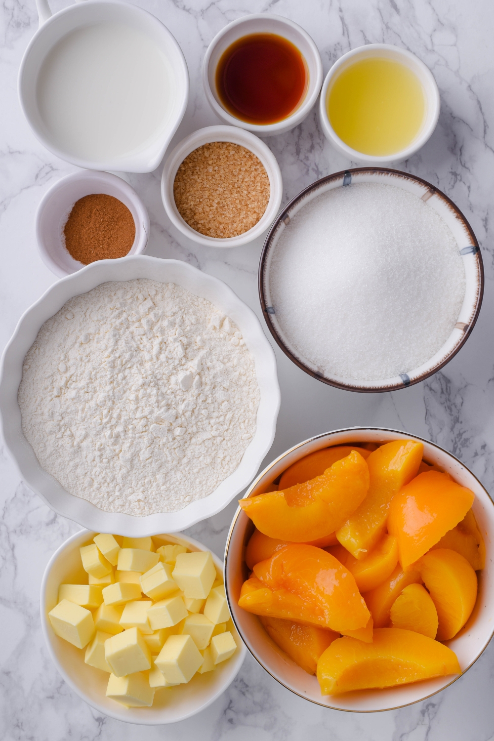 A countertop with sliced canned peaches, cubes of butter, flour, sugar, brown sugar, cinnamon, vanilla extract, lemon juice, and milk in separate bowls.