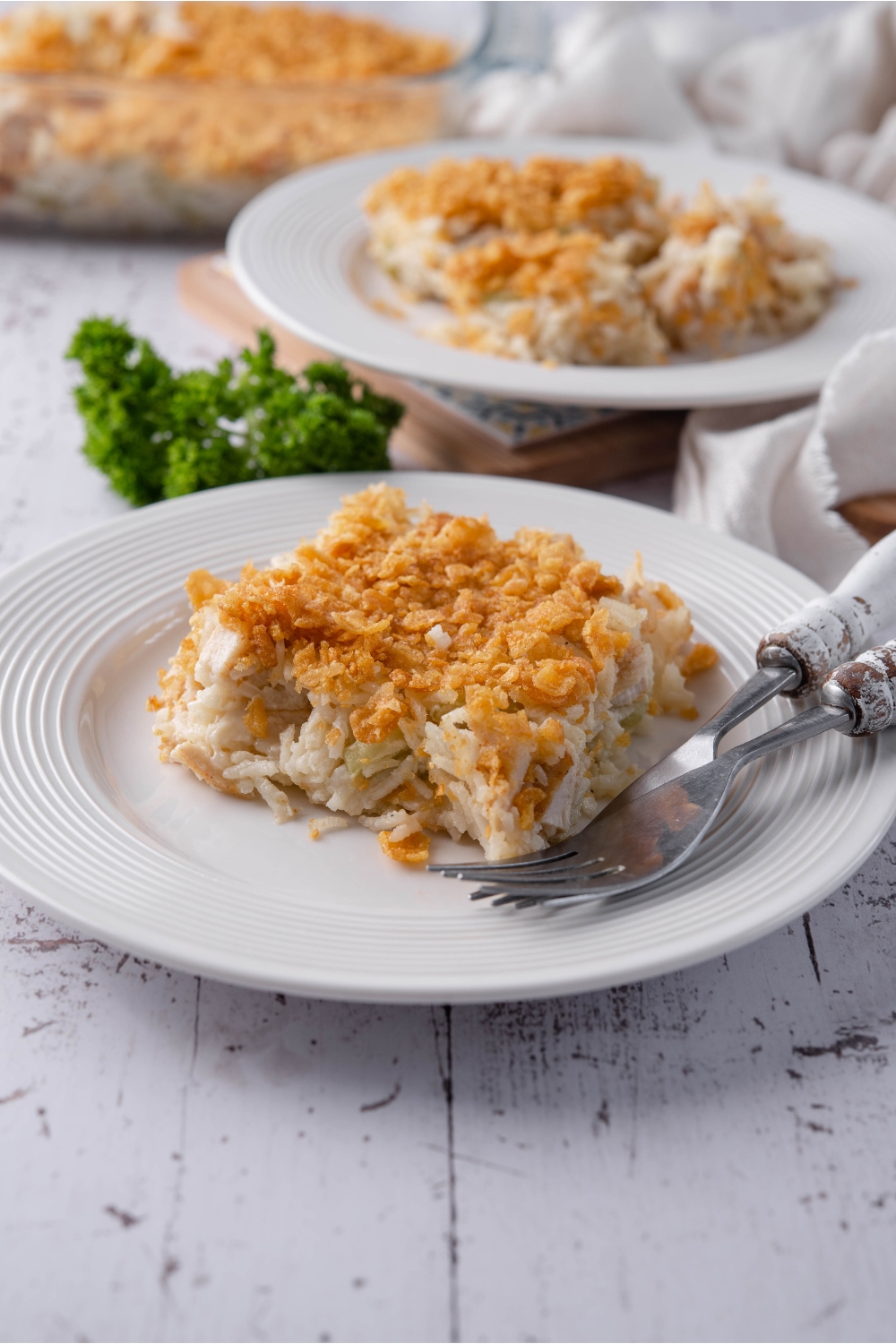 A plate with a serving of rotisserie chicken casserole topped with crushed cornflakes. There is a fork on the plate and a second serving of casserole in the background.