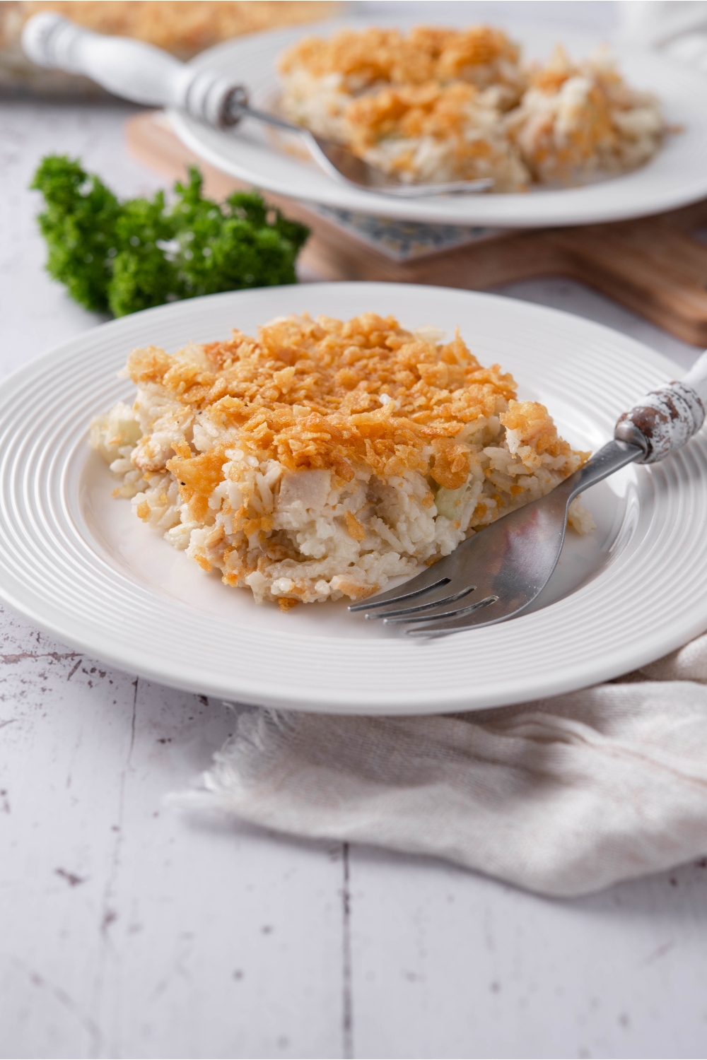 A plate with a serving of rotisserie chicken casserole topped with crushed cornflakes. There is a fork on the plate and a second serving of casserole in the background.