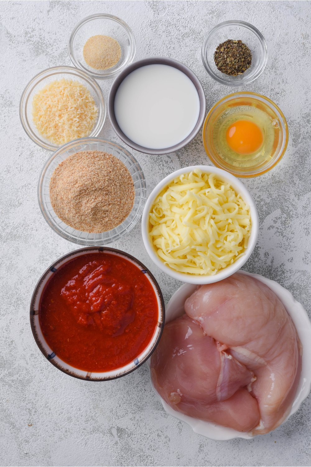 Overhead view of an assortment of ingredients including bowls of raw chicken breasts, marinara sauce, shredded cheese, an egg, breadcrumbs, seasoning, and milk.