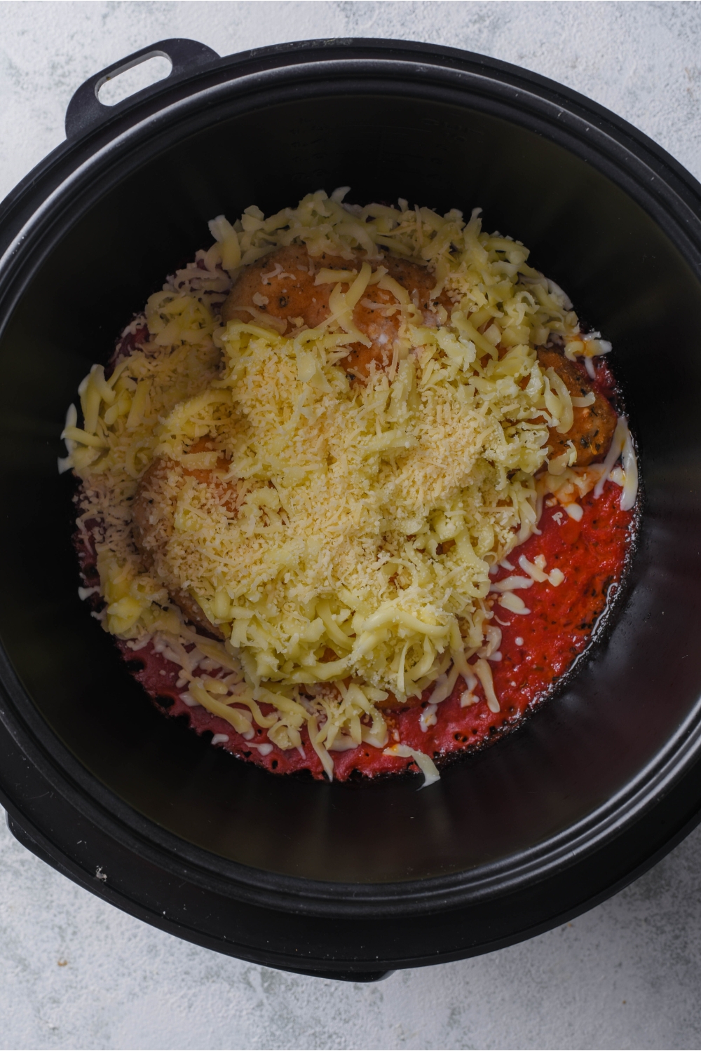 A black crockpot with two chicken breasts on top of marinara sauce and covered with shredded cheese.