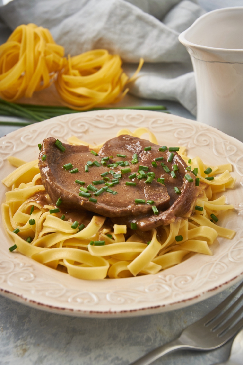 A cube steak covered in brown gravy garnished with sliced green onion and served on a bed of linguini noodles.