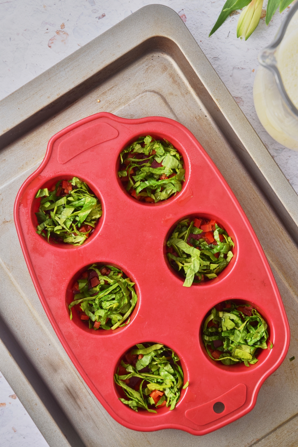 A baking sheet with a silicone muffin mold. Each mold has spinach and red peppers in it.