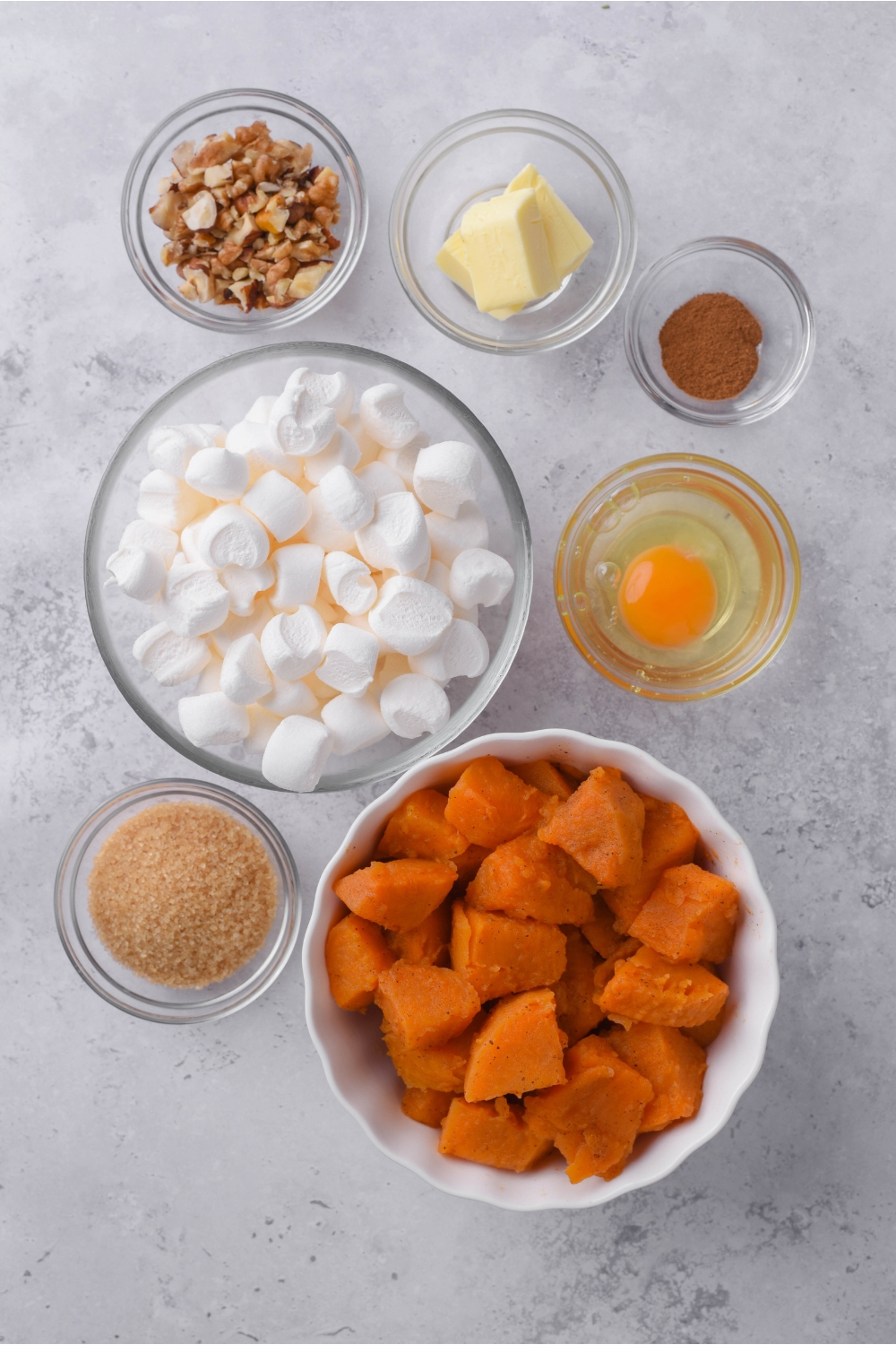 Overhead view of an assortment of ingredients including bowls of canned yams, marshmallows, an egg, brown sugar, butter, chopped nuts, and cinnamon.